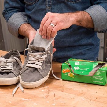 A hand places a scented dryer sheet inside a gray sneaker, with another sneaker nearby, on a wooden surface, beside a partially open Gain box.
