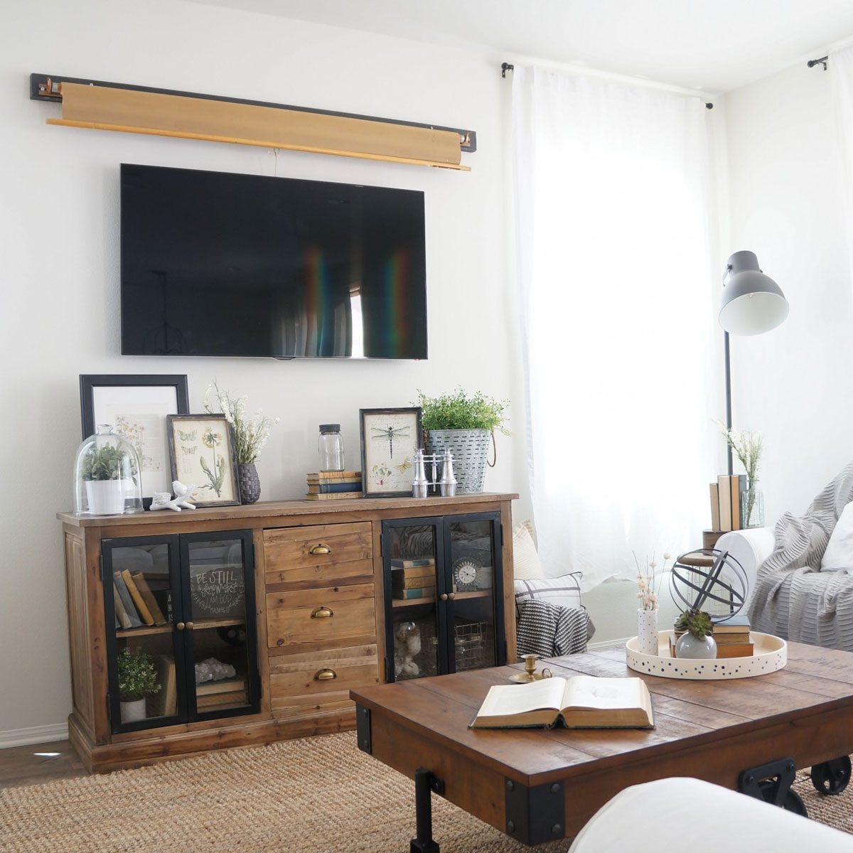 A wooden media console displays framed images and plants, while a television hangs above. Light filters through sheer curtains in a bright, cozy room.