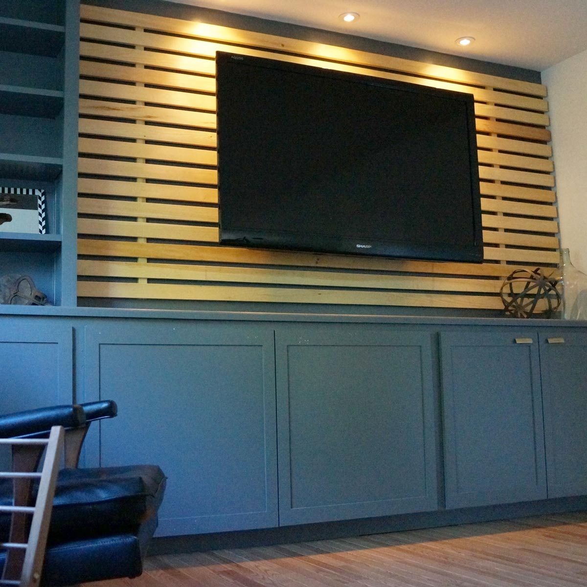 A mounted television displays against a slatted wooden wall, with soft lighting above. A dark cabinet lies below, and a black chair is nearby.