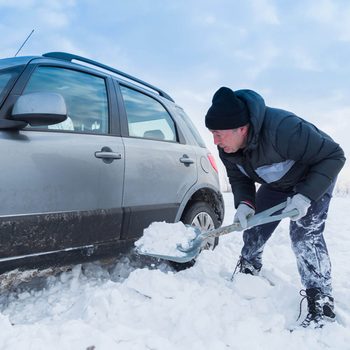 A man shovels snow from around a parked car in a snowy outdoor environment beneath a partly cloudy sky.