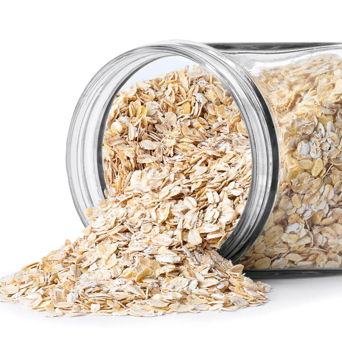 A glass jar spills rolled oats onto a white surface, creating a scattered pile beside the upright jar, suggesting a ready-to-use ingredient.