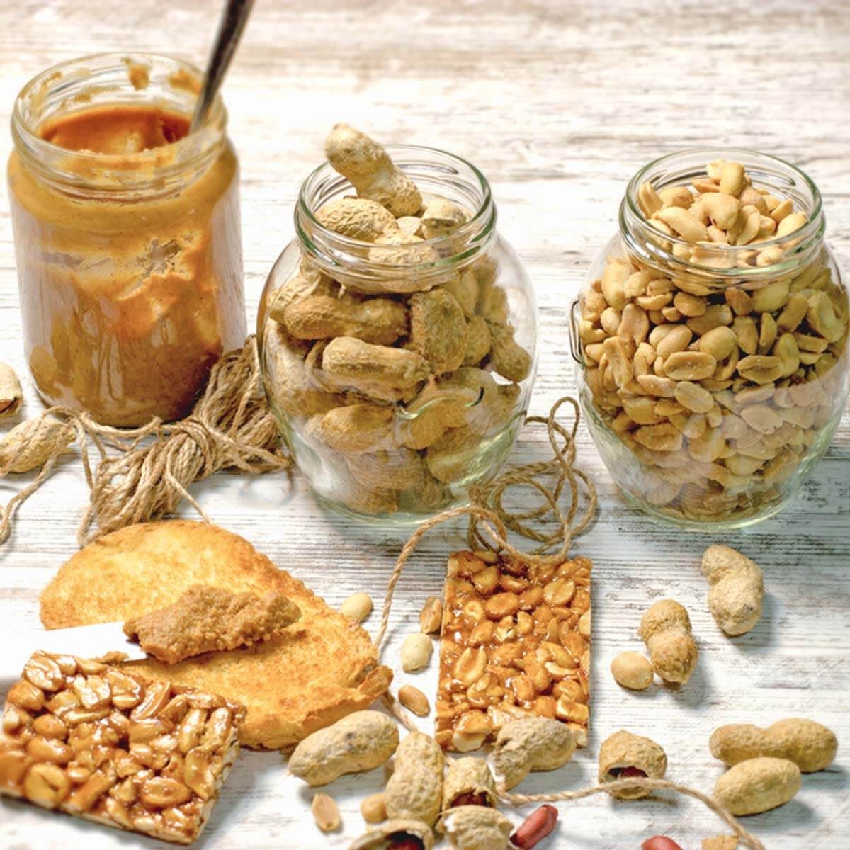Three jars containing peanuts and peanut butter sit on a wooden surface, surrounded by peanut snacks and twine, creating a cozy, natural setting.