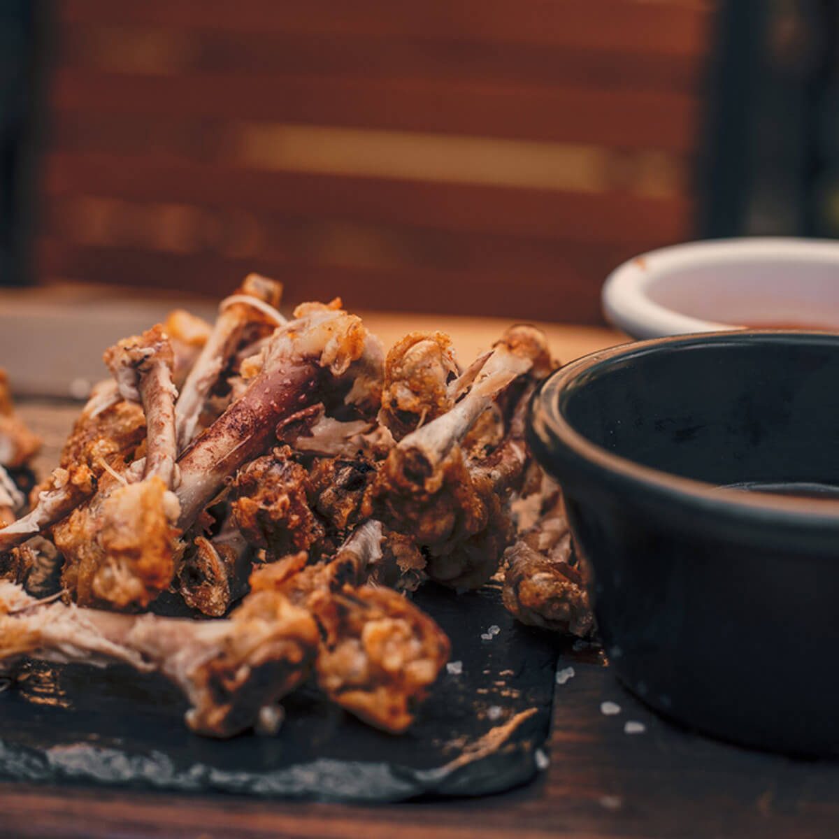 A pile of fried chicken bones rests on a dark slate plate, accompanied by a small black dish of sauce, set against a wooden background.