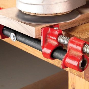 A power sander is used on a piece of wood, secured by red clamps on a workbench, highlighting a woodworking task in progress.