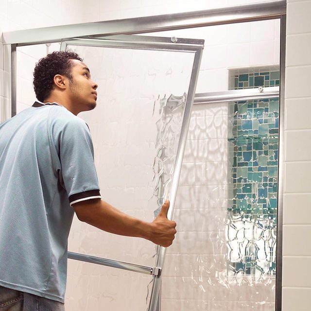 A man opens a glass shower door, revealing a tiled bathroom with white walls and a blue accent wall in the background.