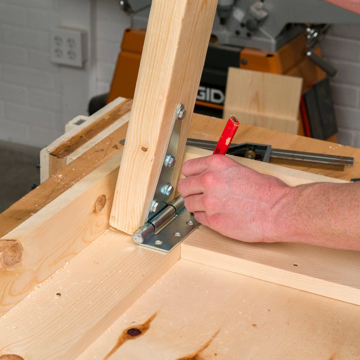 A wooden plank is being secured to a base using a metal bracket and screws, with a hand holding a red pencil for marking, in a workshop setting.