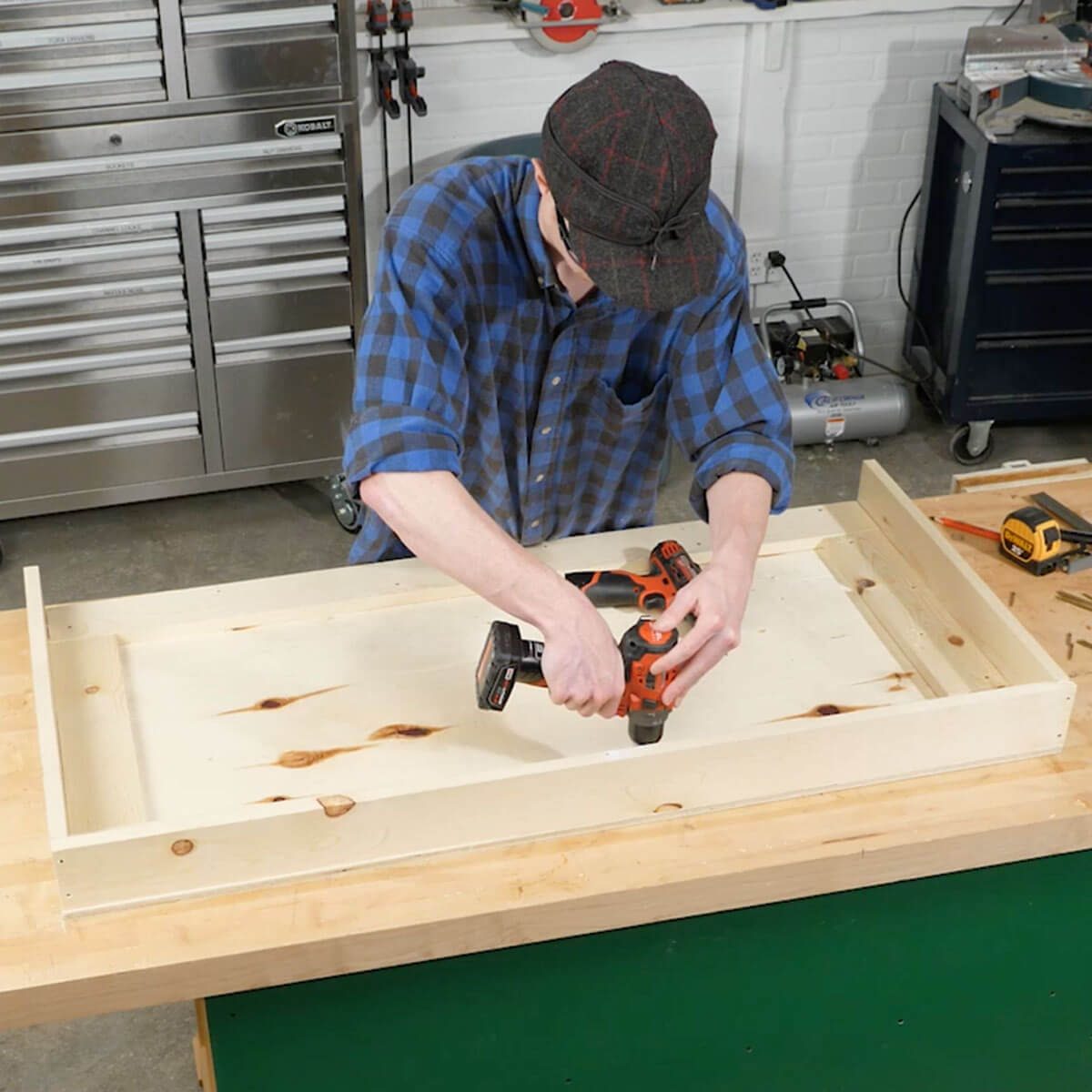 A person uses a power drill to assemble a wooden structure on a workbench in a well-lit workshop, surrounded by tools and storage units.