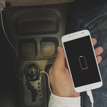 A hand holds a smartphone displaying a low battery icon, while the device is plugged into a car's power outlet amid dark interior surroundings.