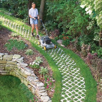 A person mows grass along a patterned stone path amidst a lush, green garden filled with various plants and surrounded by trees.