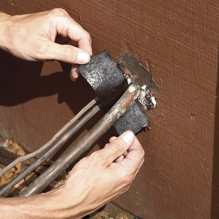 Hands apply black padding to exposed pipes near a wall, repairing or insulating them in a home maintenance context. Surrounding area shows leaves and debris.
