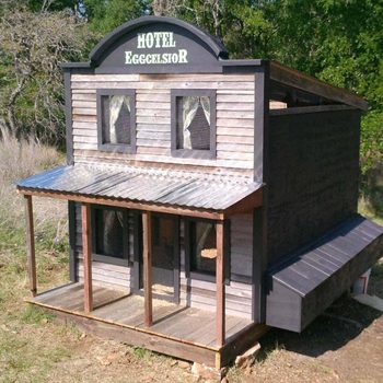 A wooden chicken coop resembling a small hotel features a sloped roof, porch, and windows with mesh. It is set in a grassy area with trees nearby.