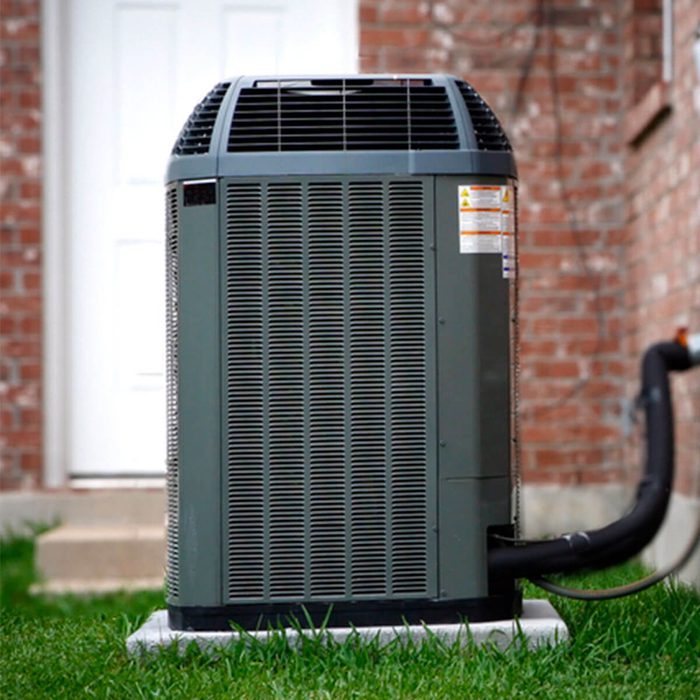 A green outdoor air conditioning unit stands on a concrete pad, connected by a black pipe, near a brick wall and grass.