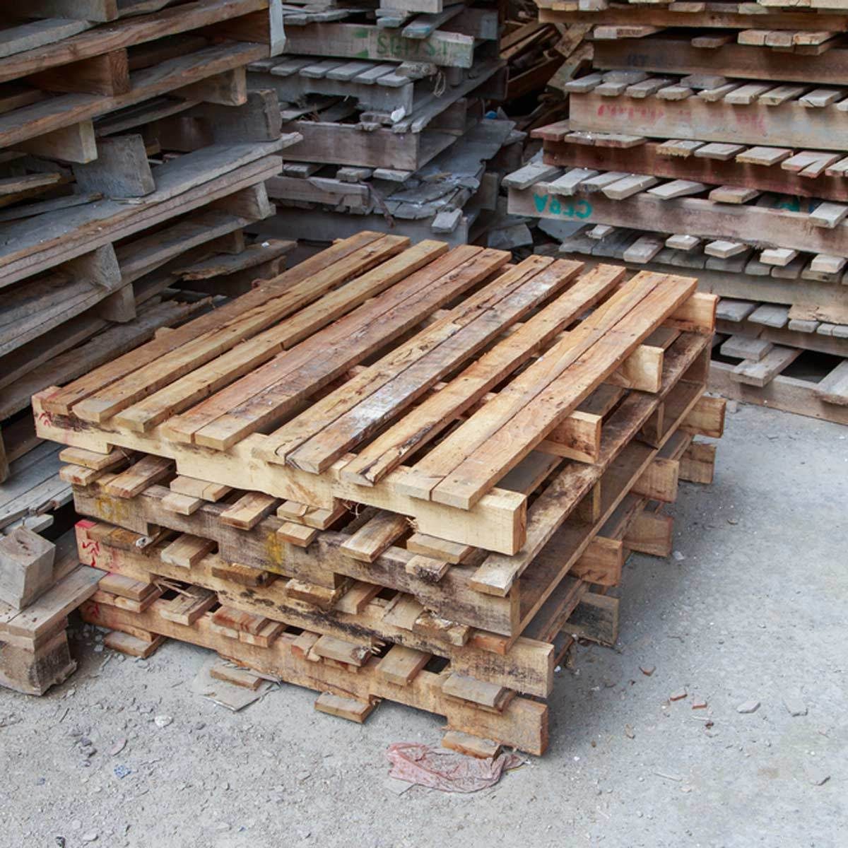 A stack of wooden pallets sits on a gravel surface, surrounded by more pallets in various orientations, indicating a storage or warehouse setting.