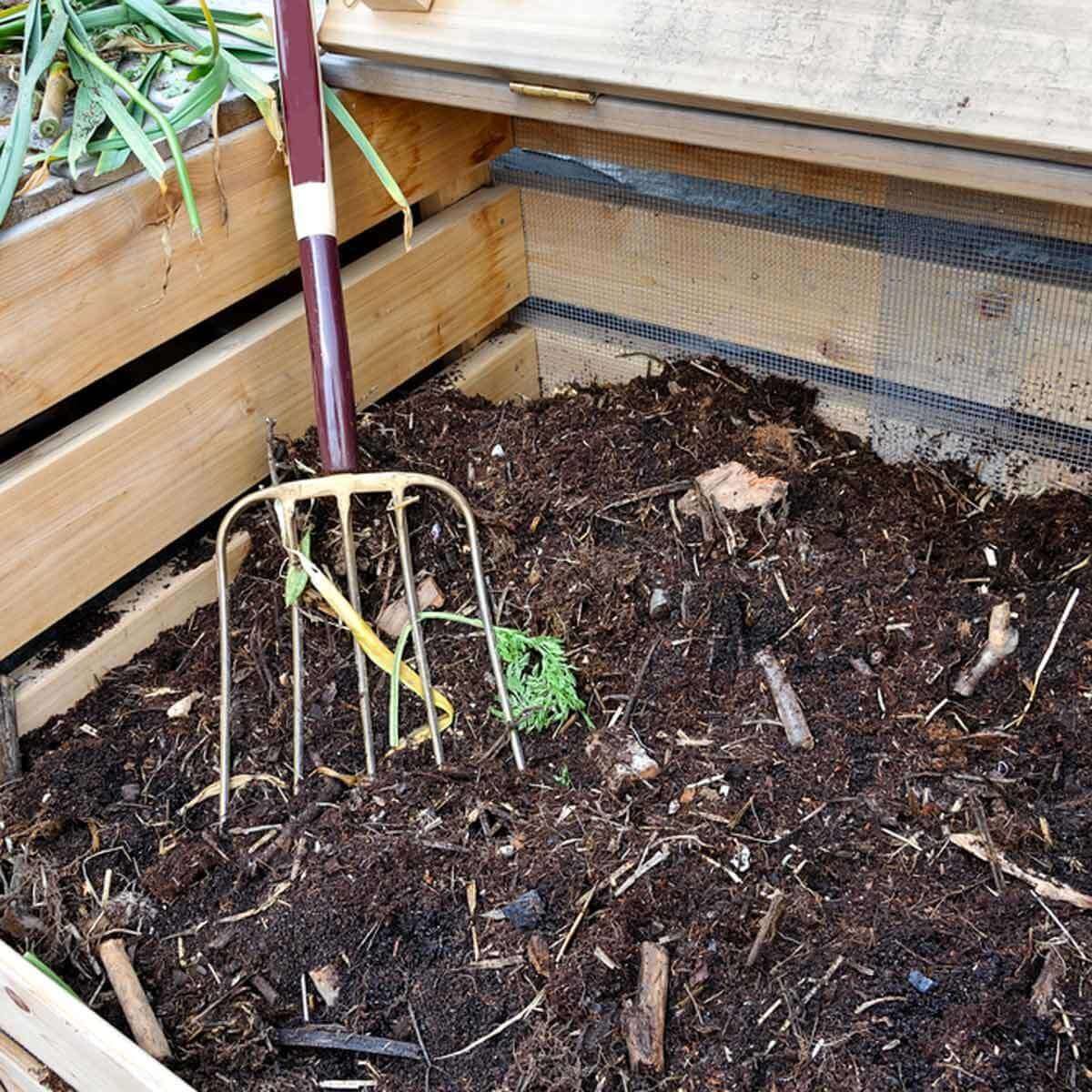 A gardening fork rests in dark, rich compost inside a wooden bin, surrounded by twigs and remnants of organic material. Green plant tops peek out.