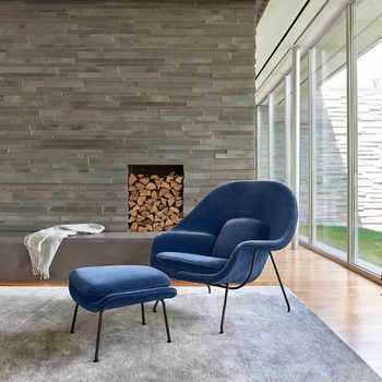 A blue chair and ottoman rest on a rug, facing a stone wall with stacked firewood. Large windows allow natural light into the room.