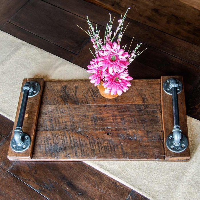 A wooden board with industrial handles supports a small vase filled with pink flowers, set on a patterned surface.
