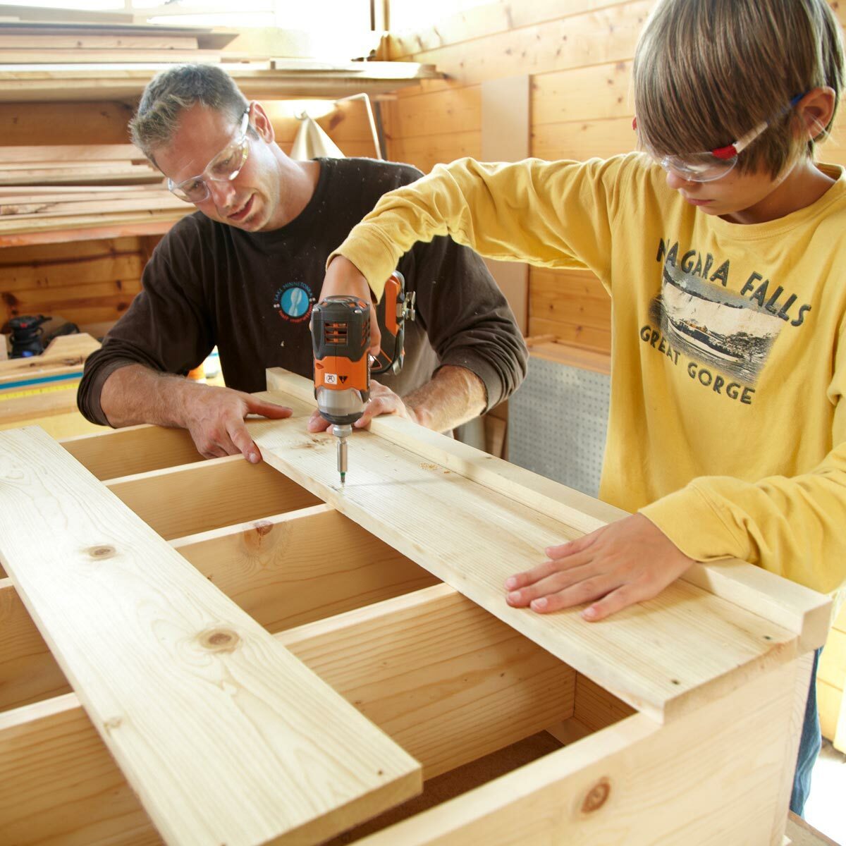 A man guides a boy drilling screws into wooden planks, within a workshop filled with tools and lumber. Sunlight illuminates the wooden interior.