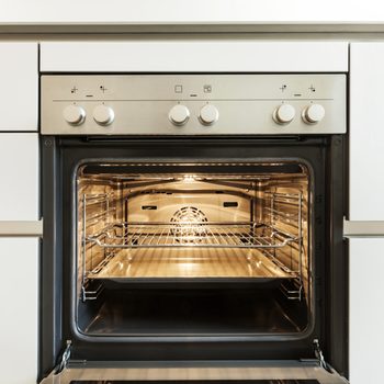 An oven with a metallic finish is open, featuring a rack and tray inside, illuminated by its internal light, set in a modern kitchen.