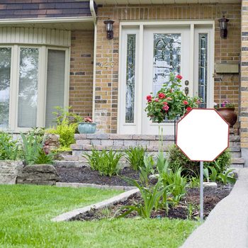 A stop sign stands beside a stone pathway leading to a home, surrounded by green grass and blooming flowers, against a backdrop of brick walls and windows.