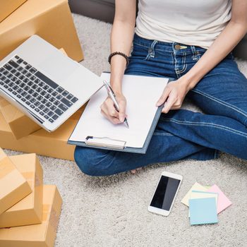 A person sits cross-legged on a carpet, writing on a clipboard with a pen, surrounded by cardboard boxes, a laptop, and sticky notes.