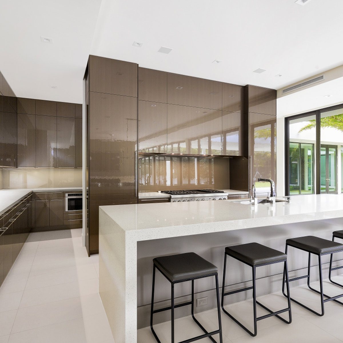A modern kitchen features sleek cabinetry, a large white countertop, and black bar stools. Natural light streams through expansive windows, enhancing the open space.