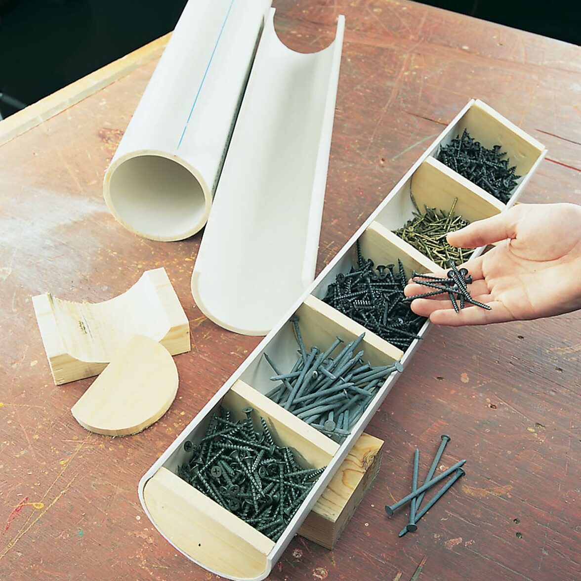 A hand holds screws from a divided wooden tray; nearby, two plastic tubes and a shaped wooden piece rest on a workbench.