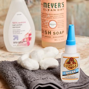 A gray towel holds cotton balls and a bottle of super glue, with nail polish remover and dish soap positioned nearby on a textured surface.