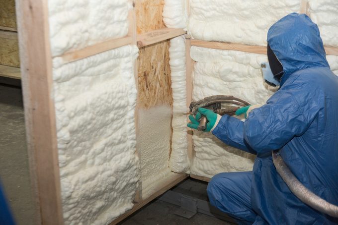 A person in protective gear sprays insulation foam into wooden wall frames in a workshop. The foam expands as it fills the cavities.
