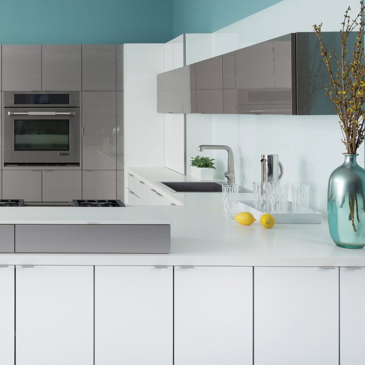 A modern kitchen features sleek cabinets and appliances, with a polished countertop displaying glassware, lemons, and a vase, surrounded by a light blue wall.