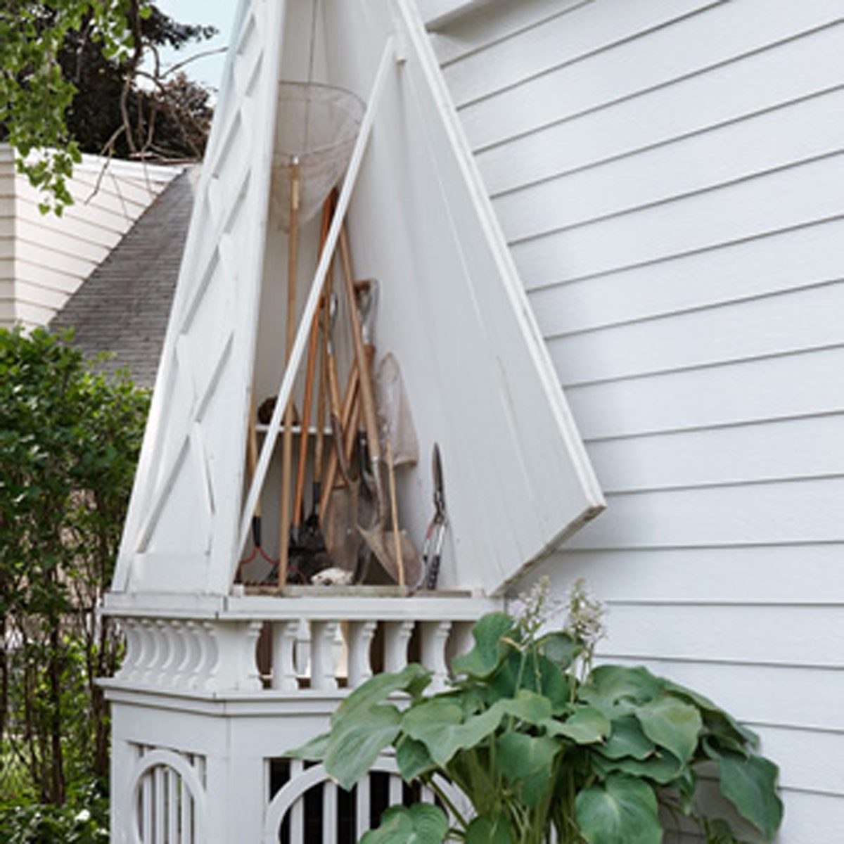 A white garden shed stores tools like shovels and a net, surrounded by plants and a white house, under a partially cloudy sky.