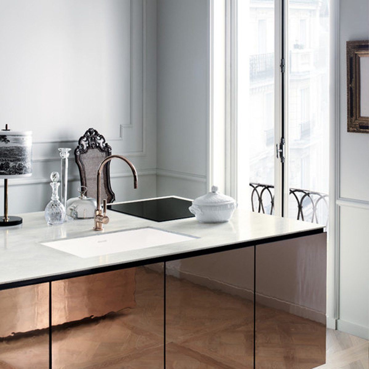 A modern kitchen features a sleek countertop with a sink and stovetop, a decorative chair, and elegant glassware beside a window providing natural light.