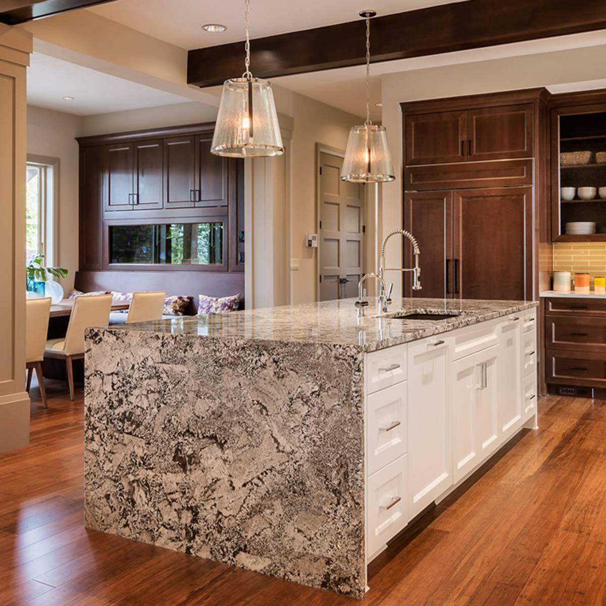 A modern kitchen features a large island with a textured stone surface, a sink, and pendant lights, surrounded by wooden cabinets and a dining area.