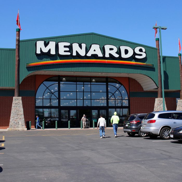 Menards store entrance features large signage as people walk toward the double glass doors on a sunny day, surrounded by parked cars.