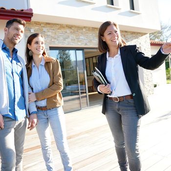 A woman gestures while showing a couple a house, standing on a wooden deck outside a modern home with large windows and a stone facade.