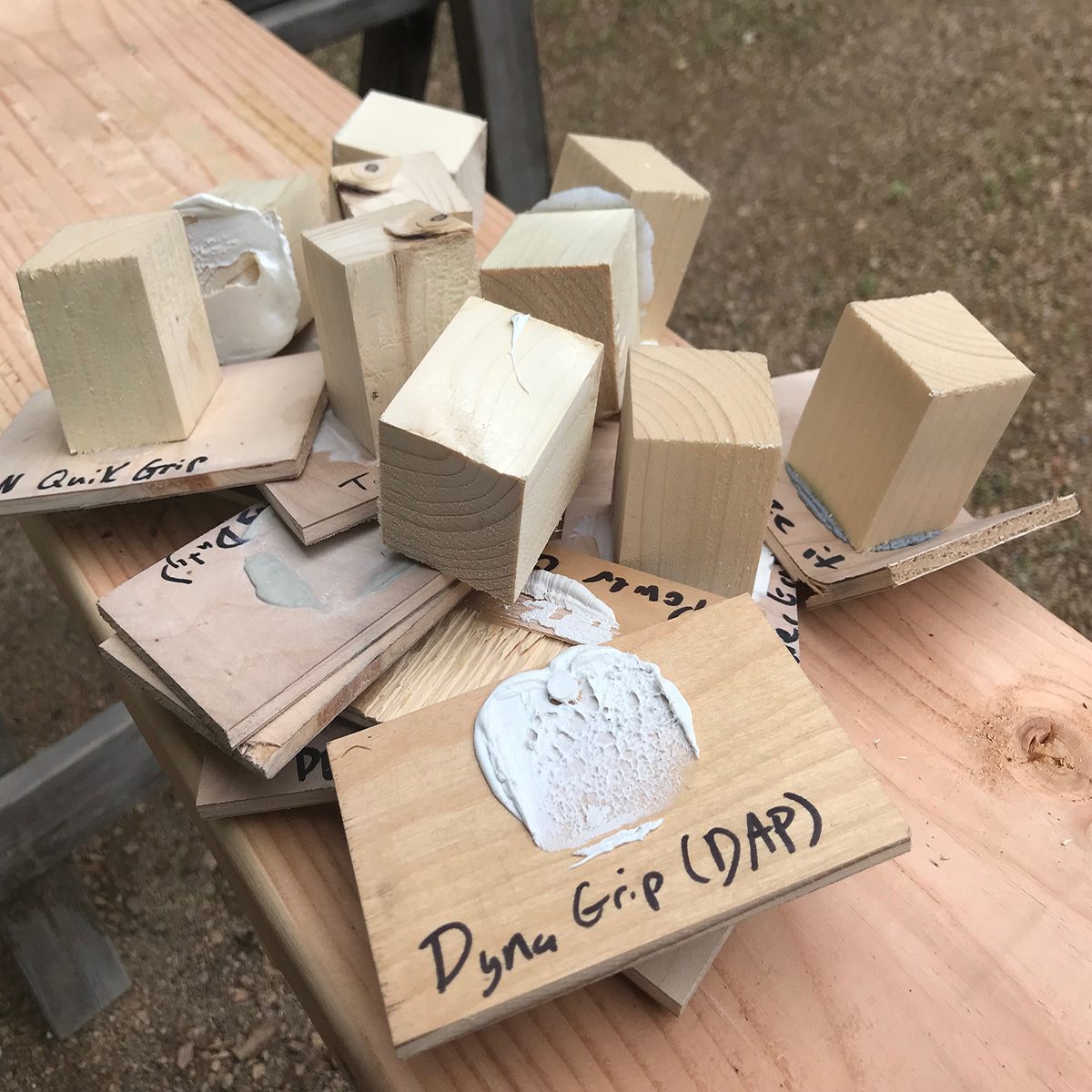 Wooden blocks are stacked on cards labeled with different glue types, resting on a light wooden surface in an outdoor setting.