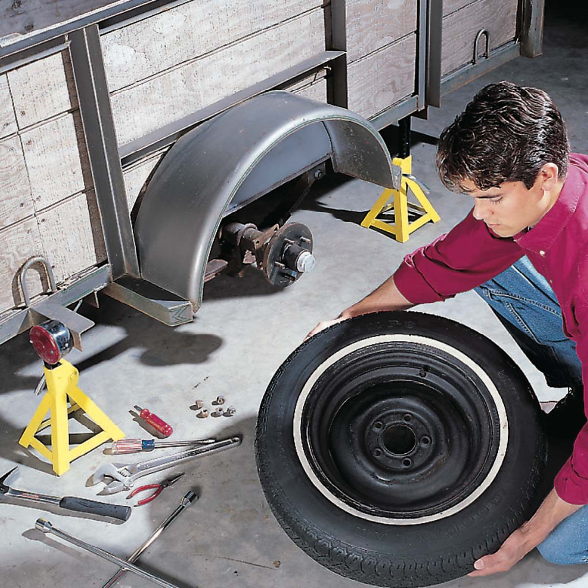 A person is holding a tire while repairing a vehicle, surrounded by tools and equipment in a garage setting, with a trailer in the background.