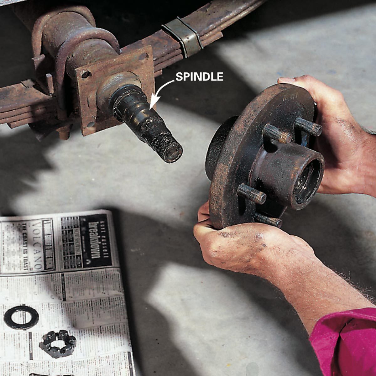 A hand holds a rusted brake hub, nearing attachment to a spindle on a vehicle axle, with tools and parts on a newspaper-covered floor. Text: "SPINDLE"