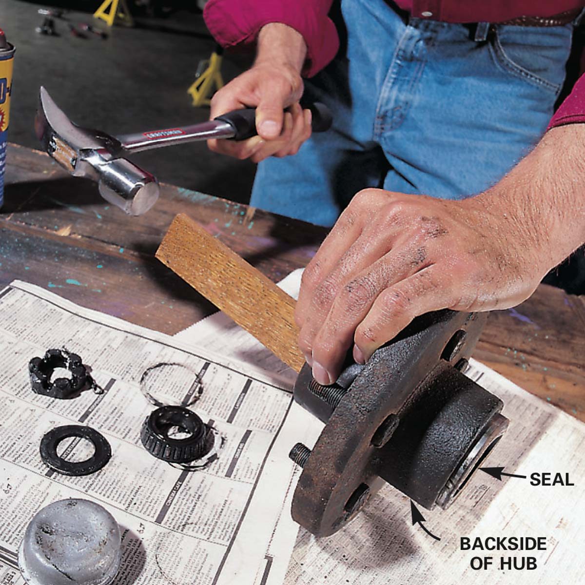 A hand grips a mechanical hub while another hand hammers a wooden block against it on a cluttered workbench, surrounded by tools and parts.