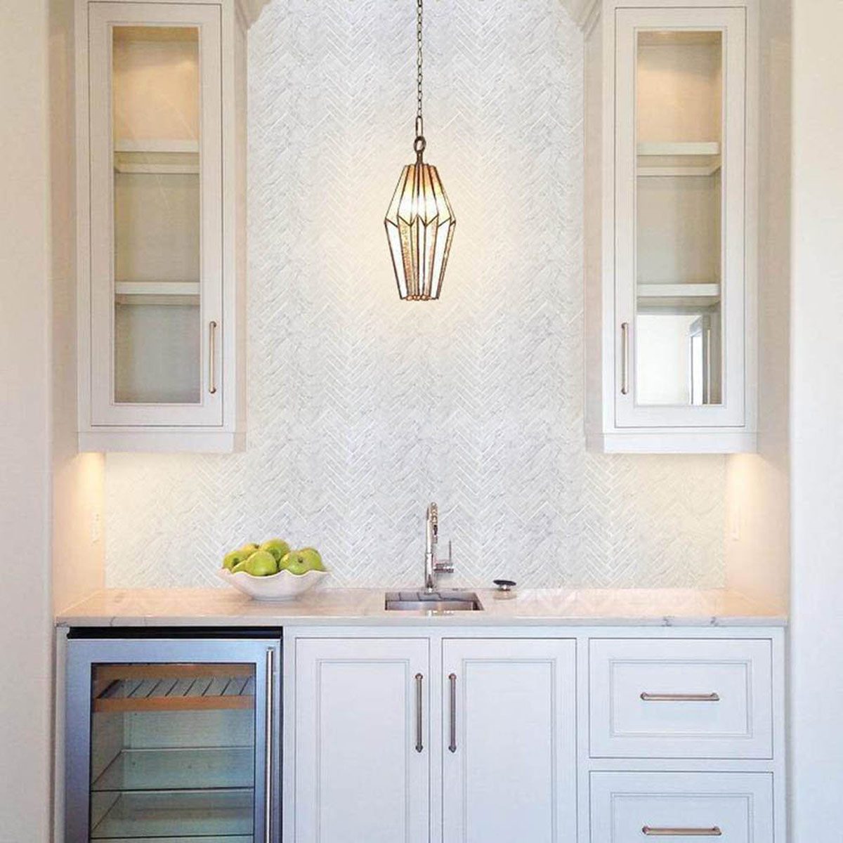 A modern kitchen corner features a sink under a stylish light fixture, with white cabinetry, a fruit bowl, and a mini fridge.