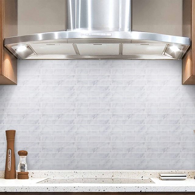A stainless steel range hood hovers over a smooth, light marble-patterned backsplash in a modern kitchen, with wooden cabinets and a granite countertop below.