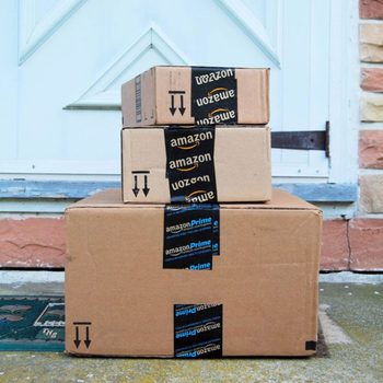 Three stacked cardboard boxes, labeled with Amazon logos, rest at a door step outside a residential building with a decorative mat nearby.