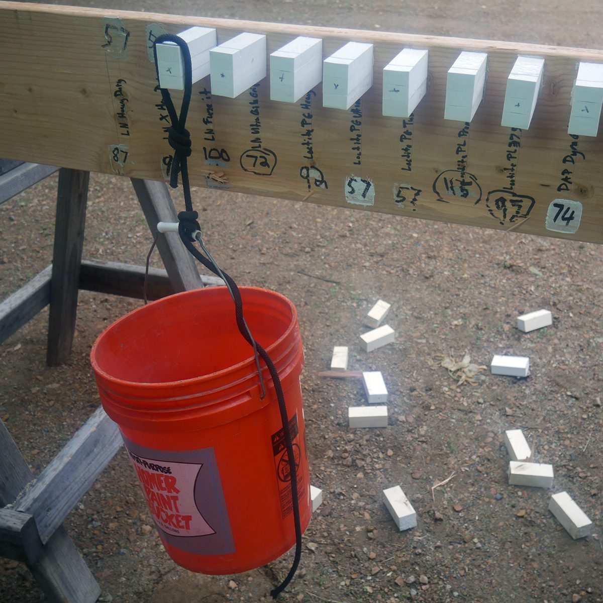 A wooden beam holds white blocks, while a bucket hangs below; some blocks are scattered on the ground. The setting appears outdoor and somewhat unfinished.