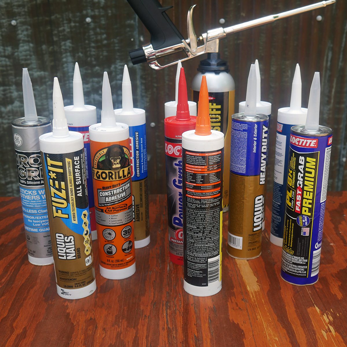Various adhesive tubes are arranged on a wooden surface, showcasing different brands and types, with a caulking gun positioned above them.