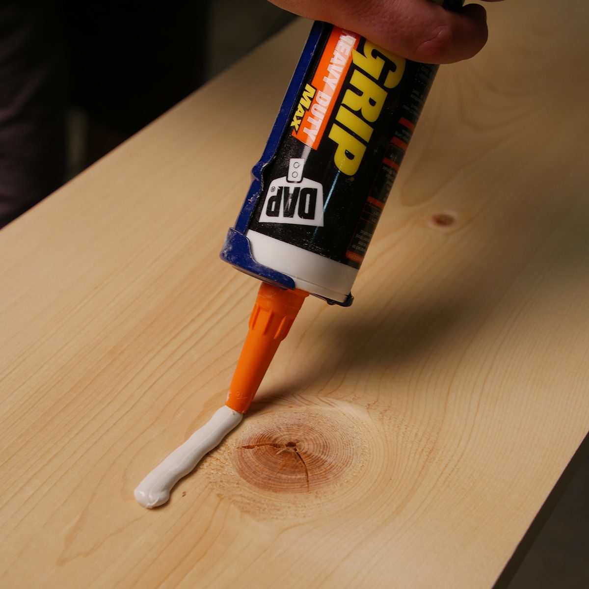 A caulking gun dispenses a white adhesive onto a wooden surface that features a prominent knot, indicating work on a project in a workshop setting.