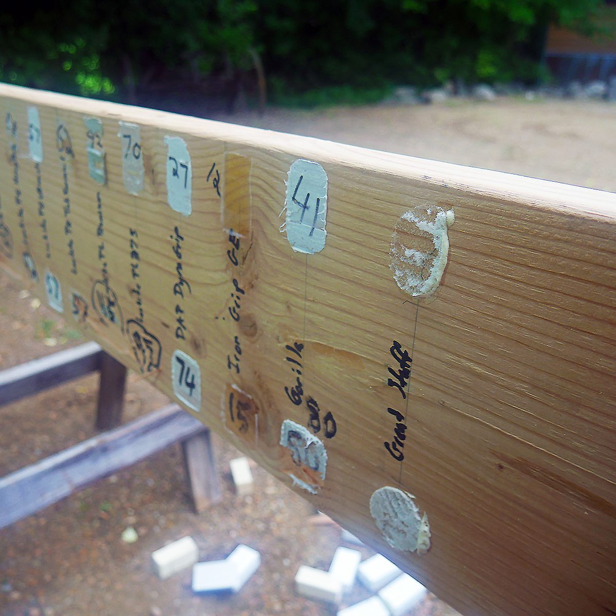 A wooden beam with label stickers and handwritten notes displays various numbers and names. It is positioned outdoors on a gravel surface surrounded by greenery.
