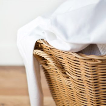 A wicker laundry basket holds white fabric, partially draped over the edge, set against a light background with wooden flooring.