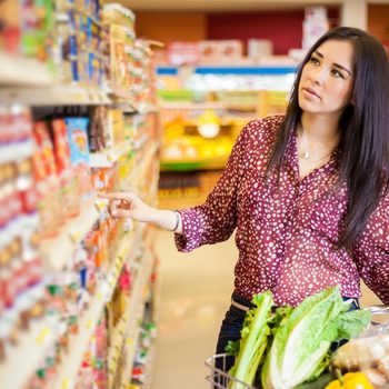 A woman reaches for a product on a grocery shelf, surrounded by colorful food packages, while a shopping cart filled with vegetables is beside her.