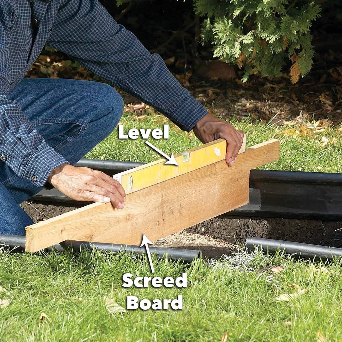 A person adjusts a level on a screed board while kneeling on grass, preparing to level a surface within a garden setting.