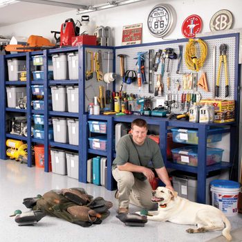 A man kneels beside a dog in a garage, surrounded by organized tools and storage bins, with hunting decoys scattered on the floor.