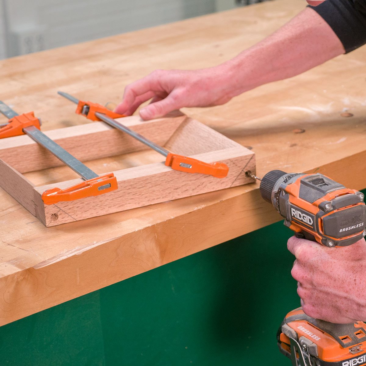 A person is using a drill on a wooden frame, secured with orange clamps, on a workbench, in a workshop setting.